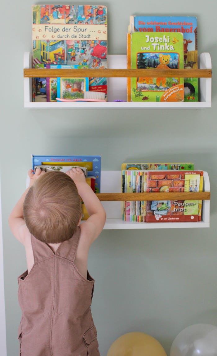 A toddler reaches for vibrant children's books on a white wall shelf, fostering early literacy.